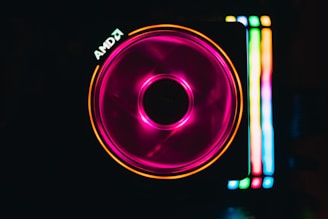 A computer cooling fan illuminated with vibrant RGB lighting. The fan features a central circular hub and spins with a pink hue, creating a mesmerizing effect. To the right, vertical strips of light display a gradient of colors, including green, blue, and yellow. The AMD logo is visible near the top left corner.