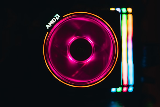 A computer cooling fan illuminated with vibrant RGB lighting. The fan features a central circular hub and spins with a pink hue, creating a mesmerizing effect. To the right, vertical strips of light display a gradient of colors, including green, blue, and yellow. The AMD logo is visible near the top left corner.