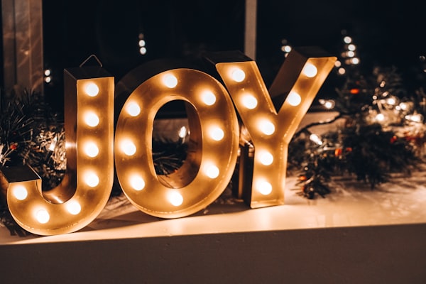 the word "joy" spelled out in lighted letters against a background of holiday lights and evergreen branches