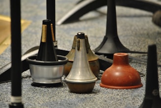 Several brass instrument mutes of different shapes and materials are arranged on a carpeted floor, surrounded by the legs of music stands.