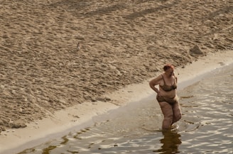 A person in a leopard print swimsuit stands in shallow water near a sandy beach. The beach stretches out with textured sand, while gentle waves approach the shore.