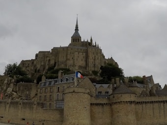 A historic and imposing stone abbey sits atop a rocky hill, characterized by its medieval architecture and tall spire. The foreground features fortified ramparts and traditional buildings made of stone with slate roofs. A French flag is visible, adding a touch of color against the cloudy sky.