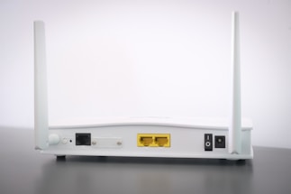 A white Wi-Fi router with two antennas positioned vertically. The router is seen from the back, showing ports and connectors including Ethernet ports, a power switch, and a power input. The device is placed on a dark surface against a light background.