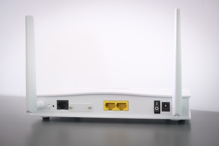 A white Wi-Fi router with two antennas positioned vertically. The router is seen from the back, showing ports and connectors including Ethernet ports, a power switch, and a power input. The device is placed on a dark surface against a light background.