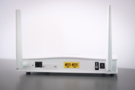 A white Wi-Fi router with two antennas positioned vertically. The router is seen from the back, showing ports and connectors including Ethernet ports, a power switch, and a power input. The device is placed on a dark surface against a light background.