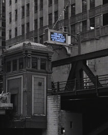 An urban scene features a tall, ornate building next to an elevated railroad or bridge. A retro-style marquee sign displays a message about an upcoming concert. The architecture appears classic and historical, with intricate detailing.