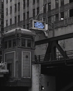 An urban scene features a tall, ornate building next to an elevated railroad or bridge. A retro-style marquee sign displays a message about an upcoming concert. The architecture appears classic and historical, with intricate detailing.