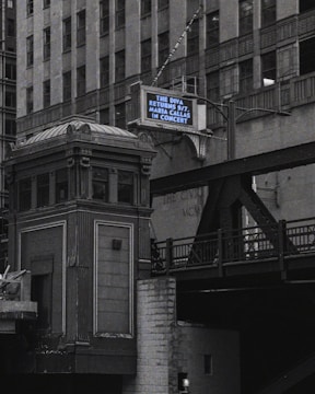 An urban scene features a tall, ornate building next to an elevated railroad or bridge. A retro-style marquee sign displays a message about an upcoming concert. The architecture appears classic and historical, with intricate detailing.