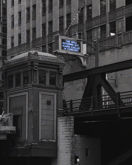 An urban scene features a tall, ornate building next to an elevated railroad or bridge. A retro-style marquee sign displays a message about an upcoming concert. The architecture appears classic and historical, with intricate detailing.