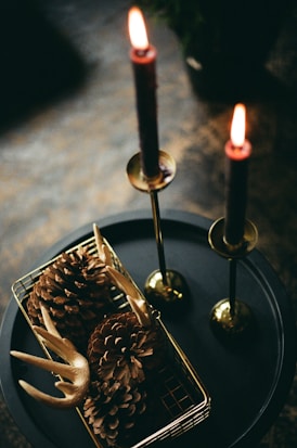 A pair of lit taper candles in tall brass candle holders sit on a black circular tray. Next to them is a small metal basket containing large pinecones and antlers, possibly intended as rustic decorations.