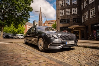 A sleek, black luxury sedan is parked on a cobblestone street, with an urban backdrop of brick buildings and a spire in the distance. The scene is filled with lush green trees and a clear blue sky, contributing to the serene atmosphere. Other vehicles and bicycles are visible, adding to the lively urban setting.