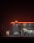 A dimly lit gas station at night with a glowing Shell logo. Two cars are parked near the gas pumps. The station has a convenience store labeled 'Shell Select,' illuminated with warm lights. The surrounding environment is dark and misty, adding a mysterious and somewhat eerie ambiance.
