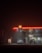A dimly lit gas station at night with a glowing Shell logo. Two cars are parked near the gas pumps. The station has a convenience store labeled 'Shell Select,' illuminated with warm lights. The surrounding environment is dark and misty, adding a mysterious and somewhat eerie ambiance.