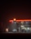 A dimly lit gas station at night with a glowing Shell logo. Two cars are parked near the gas pumps. The station has a convenience store labeled 'Shell Select,' illuminated with warm lights. The surrounding environment is dark and misty, adding a mysterious and somewhat eerie ambiance.