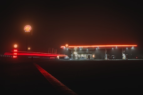 A gas station illuminated with neon red lighting, creating a vivid contrast against the dark night sky. The Shell logo is visible on signs, and lights reflect off the wet ground. Several cars are parked near the pumps, and a light trail from a moving vehicle accentuates the scene.