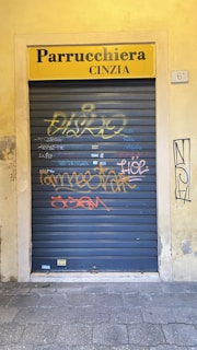 A closed metallic roller shutter with graffiti in various colors, including yellow and orange, is set within a beige wall. Above the shutter, there is a yellow sign that reads 'Parrucchiera Cinzia', indicating it's a hair salon. The ground is paved with stones, and there is additional graffiti on the surrounding wall.
