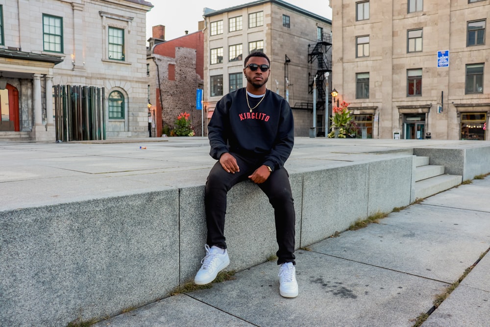 man in blue and black adidas sweater and blue denim jeans sitting on gray concrete bench