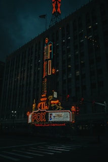 A dimly lit urban scene featuring a historic theater building with an illuminated marquee displaying the word 'FOX' in bright red neon lights. The signage includes a decorative element with a horse and a vertical sign running up the building's facade. The architecture shows gothic details and multiple windows characteristic of a theater. The American flag is visible on top of the building.