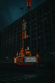 A dimly lit urban scene featuring a historic theater building with an illuminated marquee displaying the word 'FOX' in bright red neon lights. The signage includes a decorative element with a horse and a vertical sign running up the building's facade. The architecture shows gothic details and multiple windows characteristic of a theater. The American flag is visible on top of the building.