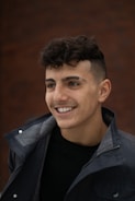 A young man with curly brown hair and a smile, wearing a dark jacket over a black shirt. The background is a blurred brown wall, giving a warm ambiance.