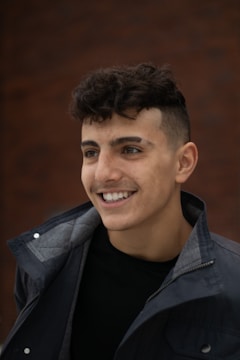 A young man with curly brown hair and a smile, wearing a dark jacket over a black shirt. The background is a blurred brown wall, giving a warm ambiance.