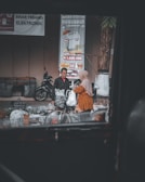 A street scene featuring two people standing beside a bicycle laden with bags in an outdoor market setting. The focus is on the interaction between the two individuals, one wearing a headscarf and an orange patterned garment, and the other in a dark jacket. There are several electronic advertisement posters in the background, along with parked motorcycles and various baskets and goods on the ground.