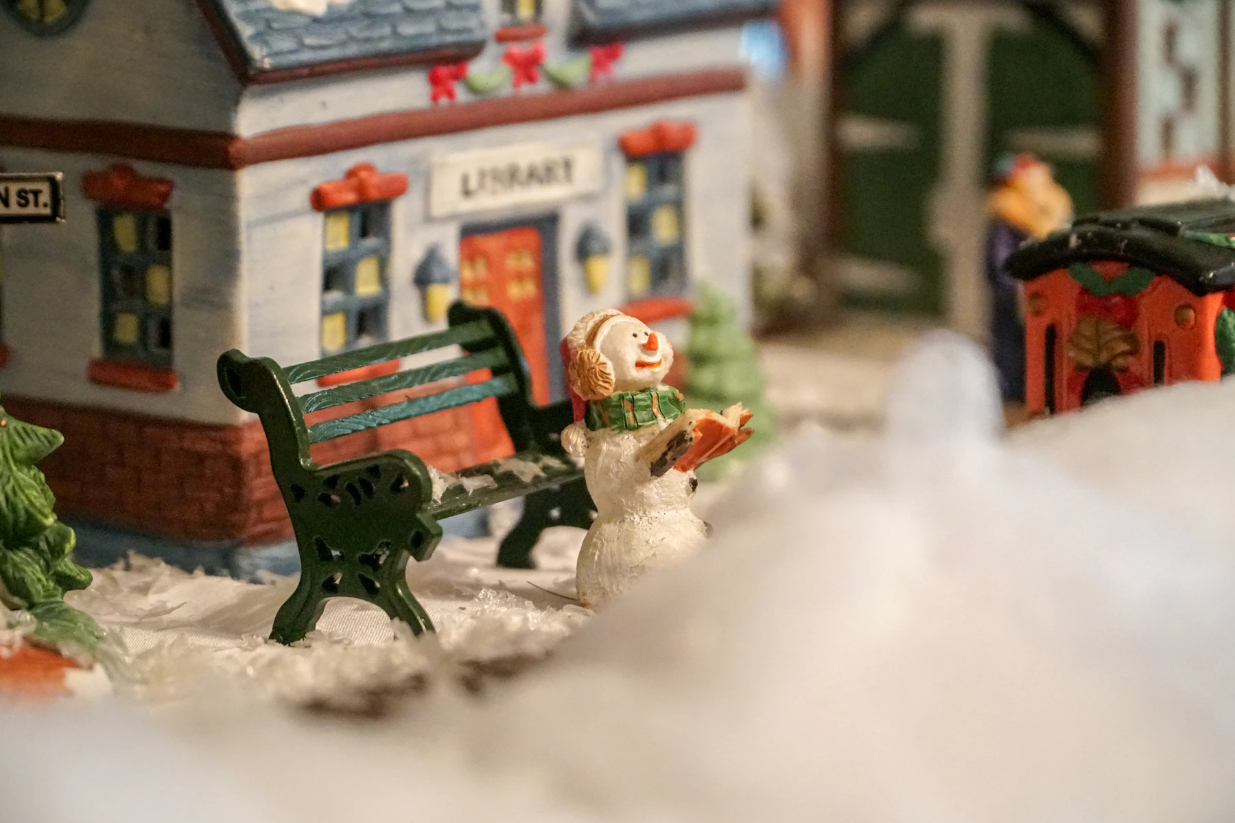 Close-up of a snowman figure reading on a green bench in front of a painted village library