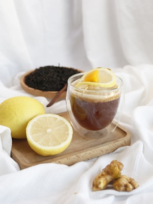 A glass of dark tea garnished with lemon slices on a wooden tray surrounded by whole and sliced lemons, ginger root, and loose tea leaves in a bowl. The setting has a clean, minimalistic look with a white fabric background.