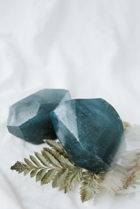 Two large, faceted dark teal crystals are placed on a light background alongside a fern leaf. The crystals have a glossy texture, and the leaf adds a natural element to the composition.
