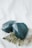 Two large, faceted dark teal crystals are placed on a light background alongside a fern leaf. The crystals have a glossy texture, and the leaf adds a natural element to the composition.