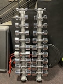 Rows of shiny chrome dumbbells are neatly arranged on two vertical racks in a gym setting. The background features a black wall with a perforated metal stair railing on the left side.