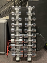 Rows of shiny chrome dumbbells are neatly arranged on two vertical racks in a gym setting. The background features a black wall with a perforated metal stair railing on the left side.