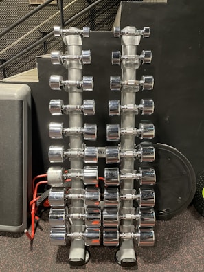 Rows of shiny chrome dumbbells are neatly arranged on two vertical racks in a gym setting. The background features a black wall with a perforated metal stair railing on the left side.