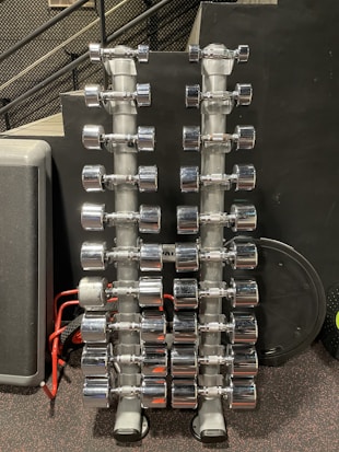 Rows of shiny chrome dumbbells are neatly arranged on two vertical racks in a gym setting. The background features a black wall with a perforated metal stair railing on the left side.