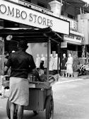 A street vendor pushes a cart in front of a store with mannequins displaying clothing. The store signs are prominently visible, with people casually walking in the background.