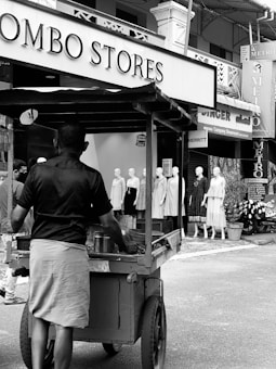 A street vendor pushes a cart in front of a store with mannequins displaying clothing. The store signs are prominently visible, with people casually walking in the background.