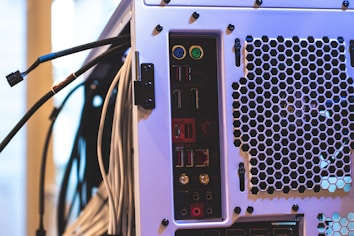 The image features the back panel of a computer tower with various ports and connectors, including USB ports, audio jacks, and PS/2 connectors, alongside a ventilation area with a hexagonal mesh design. Numerous cables are connected to the ports and extend outward.
