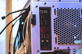 The image features the back panel of a computer tower with various ports and connectors, including USB ports, audio jacks, and PS/2 connectors, alongside a ventilation area with a hexagonal mesh design. Numerous cables are connected to the ports and extend outward.