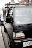 A small black truck parked on the side of a street with a visible license plate from Ontario. Buildings are visible in the background. The truck appears to be in an urban setting, and another car is partially visible next to it.