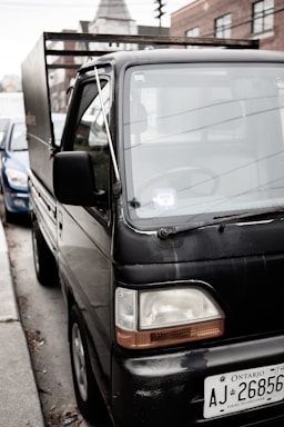 A small black truck parked on the side of a street with a visible license plate from Ontario. Buildings are visible in the background. The truck appears to be in an urban setting, and another car is partially visible next to it.