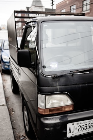 A small black truck parked on the side of a street with a visible license plate from Ontario. Buildings are visible in the background. The truck appears to be in an urban setting, and another car is partially visible next to it.