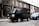 A black mini truck is parked on a street lined with brick residential buildings. There is a blue car parked closely behind it. The street appears quiet with bare trees and a gray sidewalk. The truck has some text in an Asian script on its side.