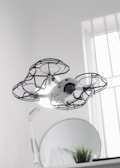 A drone hovers indoors, enclosed in a protective cage around its propellers, providing an added layer of safety. Behind it, a round mirror is visible next to a small plant, and a softly lit window allows natural light to filter into the room, creating a clean and modern aesthetic.