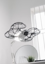 A drone hovers indoors, enclosed in a protective cage around its propellers, providing an added layer of safety. Behind it, a round mirror is visible next to a small plant, and a softly lit window allows natural light to filter into the room, creating a clean and modern aesthetic.