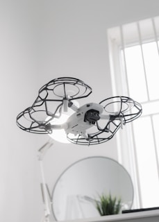 A drone hovers indoors, enclosed in a protective cage around its propellers, providing an added layer of safety. Behind it, a round mirror is visible next to a small plant, and a softly lit window allows natural light to filter into the room, creating a clean and modern aesthetic.