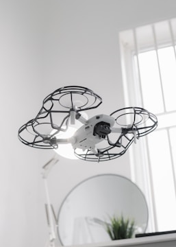A drone hovers indoors, enclosed in a protective cage around its propellers, providing an added layer of safety. Behind it, a round mirror is visible next to a small plant, and a softly lit window allows natural light to filter into the room, creating a clean and modern aesthetic.