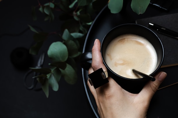 Close-up of hands holding a cup of tea next to a notebook and glasses on a wooden table.