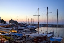 A calm harbor scene with several sailboats and yachts docked, softly lit by the fading evening light. The background shows a clear sky transitioning from warm hues of yellow and orange near the horizon to cooler blues higher up. On the left, a row of buildings with outdoor seating suggests a coastal town setting.