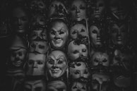 A collection of theatrical masks arranged closely together, featuring a variety of expressions and designs. The masks are predominantly monochrome, suggesting a dramatic or mysterious atmosphere.