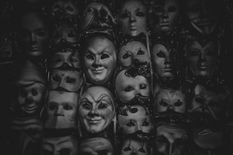 A collection of theatrical masks arranged closely together, featuring a variety of expressions and designs. The masks are predominantly monochrome, suggesting a dramatic or mysterious atmosphere.